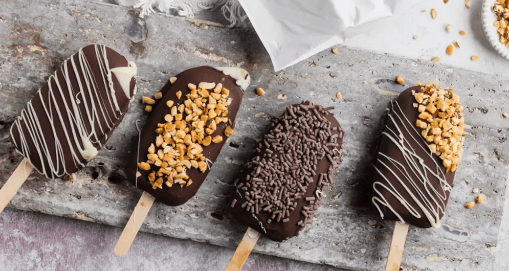 Four chocolate-dipped ice cream bars with different toppings on a stone surface.