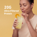 A woman drinking from a tall glass of pale yellow protein drink with a straw, garnished with an orange slice, against a warm yellow background. White text on the left reads '20G Ultra-Filtered Protein'.