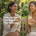 Two women laughing on a wooden bench outdoors, each holding a tall glass of iced drink with a straw. White text across the left side reads, 'Clear Protein That Supports Your Hydration.