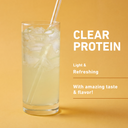 A tall clear glass filled with a pale yellow iced drink and a clear straw, set against a warm yellow background with white text reading 'CLEAR PROTEIN' and additional descriptive copy.