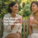 Two women sitting outdoors, holding pink drinks with straws, surrounded by greenery.