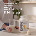 A person in a white blouse stands at a kitchen counter holding a glass of milk beside a large tub of Isopure Zero Carb Protein powder and a scoop of powder. White text on the image reads, 'Supports Health and Wellness with 22 Vitamins & Minerals.